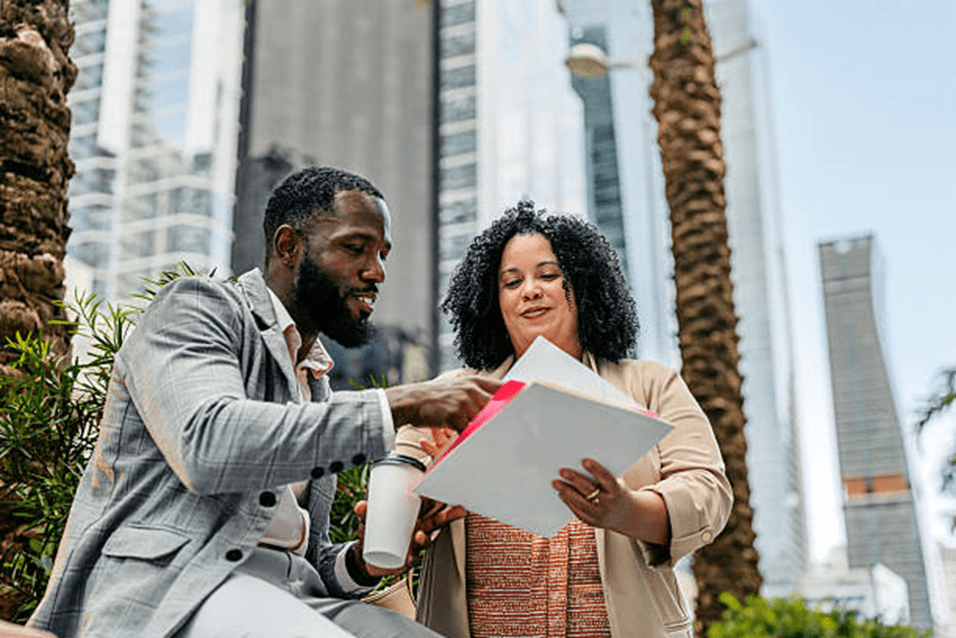Two professionals discuss documents outdoors in a city setting.